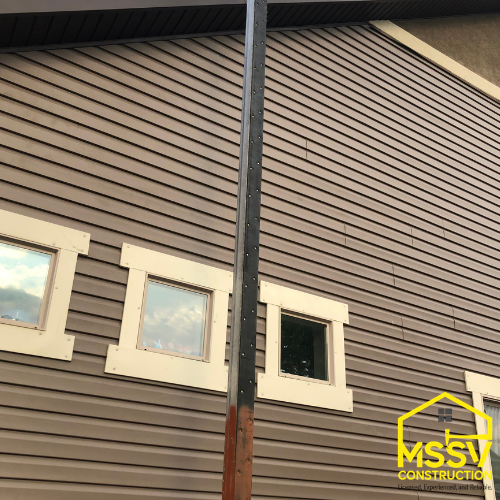 Dark brown horizontal siding with three white trimmed windows and exterior downspout.